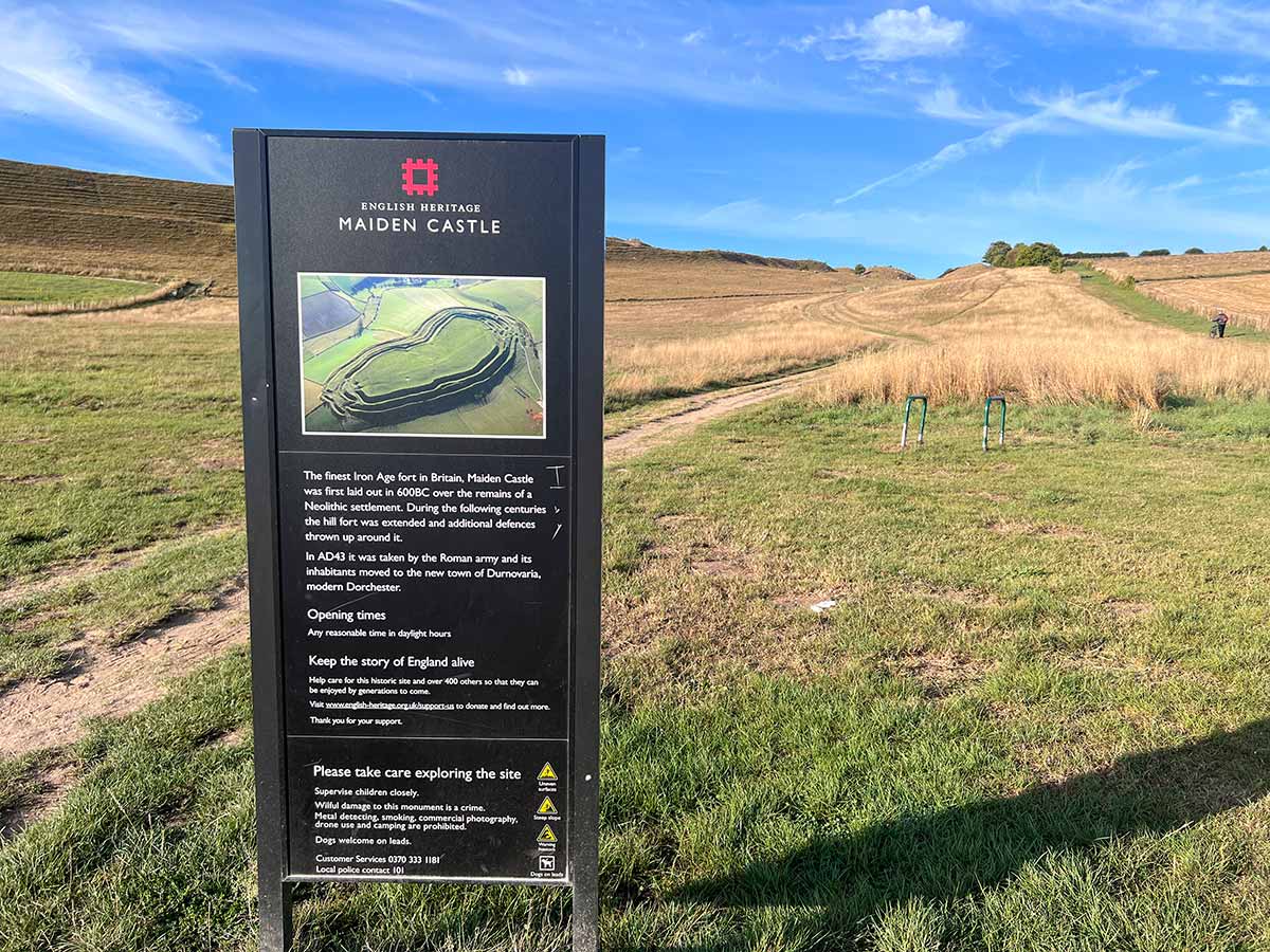 Information board about Maiden Castle by English Heritage with path towards the hill fort leading up in the background